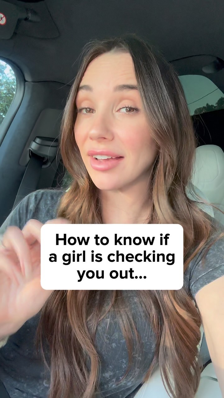 A woman is seated in a car, directly facing the camera, and is wearing a gray t-shirt. The background is blurred, with a white text graphic over her and the car interior visible. She has brown hair and is lightly smiling as she gives speaking guidance.