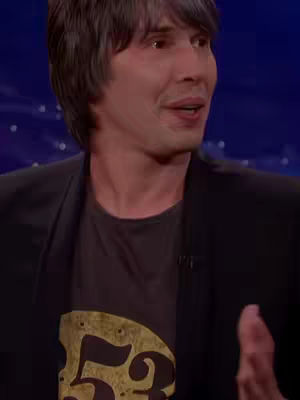 A medium shot shows a man speaking, set against a blue backdrop. The man, who appears to be in his 40s or 50s, is wearing a black blazer over a graphic t-shirt. His dark hair is neatly styled, and he appears engaged in conversation.