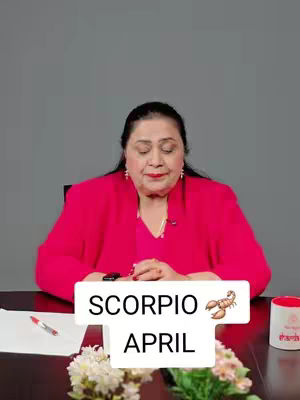 A woman in a pink blazer is seated at a table. The image has a text overlay with the words "SCORPIO" over the word "APRIL". A small potted plant with flowers and a cup with text is in the image.