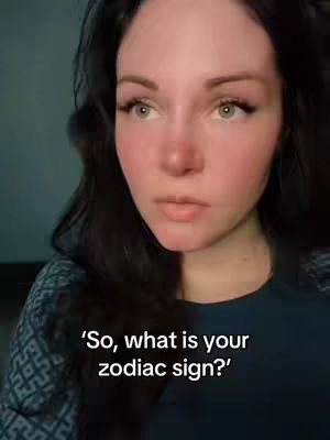 A close-up shot features a fair-skinned woman with long, dark hair. She has light eyes, and her cheeks appear to be flushed. She has applied makeup, including mascara and lipstick. The text overlayed says, “So, what is your zodiac sign?”, indicating that the video will likely address astrology or zodiac signs.