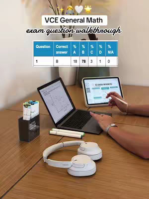 A well-lit, top-down shot of a table setup for studying. A laptop displaying exam questions, along with a table showing a laptop, a tablet with a similar interface, a notebook, headphones, and a collection of colorful markers. The image conveys a studious and organized environment.