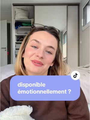 A woman with blonde hair is centered in the frame, looking towards the camera. She is wearing a brown sweater. Behind her is a bedroom setting, with a closet and a glimpse of a bed. A text overlay in a blue rounded rectangle reads: "disponible émotionnellement ?". A Tik Tok logo is in the top right.