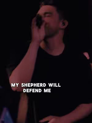 A man in a dark-colored shirt is singing into a microphone, possibly on a stage. Text overlayed on image.