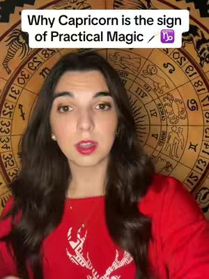 A woman with long, dark brown hair is centered in the frame, looking directly at the viewer. She is wearing a red long-sleeved shirt and is set against a backdrop of a circular zodiac wheel and an ornate tapestry. Above her head is a rectangular white banner with the text 'Why Capricorn is the sign of Practical Magic / ♑'. The overall composition is well-lit and focused on the woman, suggesting a personal address to the audience.