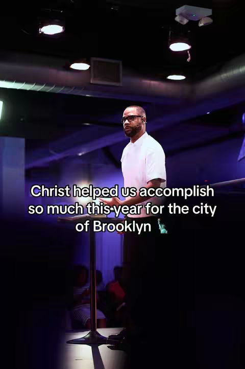 The image shows a man wearing glasses and speaking from behind a podium in a church. The background reveals a modern interior with lighting fixtures, presumably in Brooklyn.