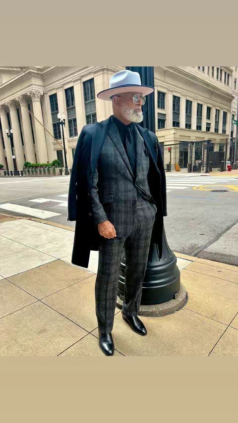 A well-dressed man stands on a city sidewalk, posing confidently. He wears a stylish gray suit, a black shirt, and a fedora. The background shows a classic building and a partially visible street, suggesting an urban setting, and adds to the image's composed and stylish aesthetic.