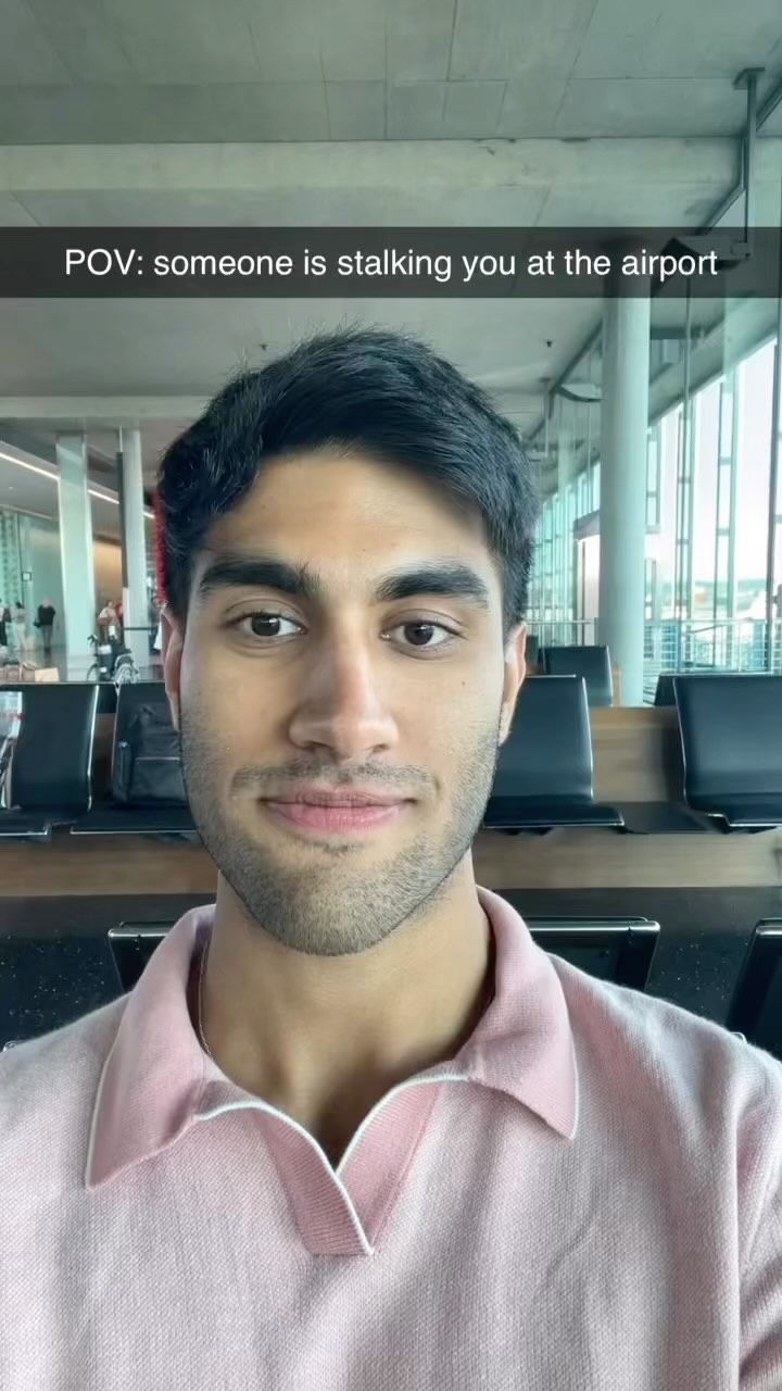 A selfie-style shot features a young man in a modern airport setting. He is framed from the chest up, centered with a neutral expression, wearing a pink polo shirt with a white collar. Behind him, a blurred backdrop of the airport terminal reveals seating, windows, and a glimpse of other people, suggesting a public, travel-related environment.