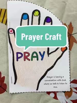 The image centers on a white cut-out of a hand showing a craft idea. Overlayed on the hand is a light teal speech bubble with white text that reads, 'Prayer Craft'. The letters P-R-A-Y are written on the fingers in multi-colored lettering. The nails are painted in different colors. Near the bottom, there's a small paragraph explaining prayer as a conversation with God. The background appears to be a warm, patterned surface.