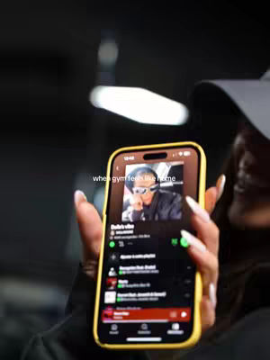 A person's hand holding a phone. The phone displays a music playlist with a photo of a person wearing sunglasses and a black jacket. The background is dark and blurred.