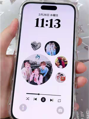 A close-up shot of a phone held in a hand, displaying a custom live wallpaper. The wallpaper features pictures of a Japanese pop group, a silver heart, and the date and time. The phone screen is framed within a clear protective case.