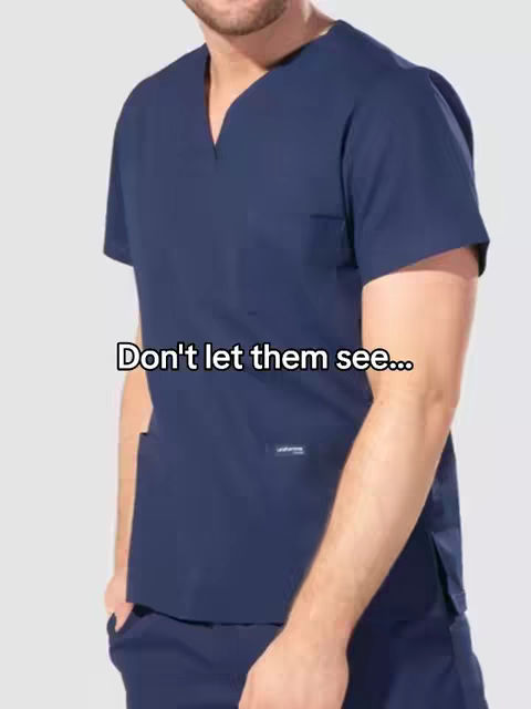 A person wearing dark blue scrubs stands against a light gray background. Text overlayed on the image reads 'Don't let them see...' The person is wearing a scrub top with a V-neck and a pocket on the chest. The person has a beard and is looking down. The angle is from the chest up.
