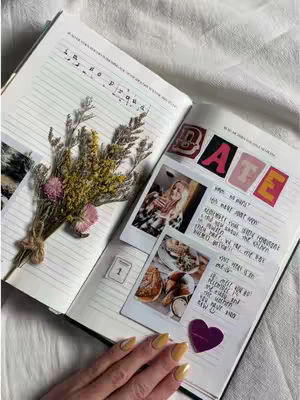 An open notebook lies on a textured white surface. The left page displays handwritten words, a small photo, and a sprig of dried flowers. The right page has photos and handwritten notes. A hand with yellow nails is in the lower part of the image.