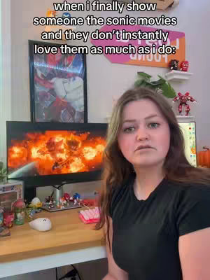 A woman with brown hair and a black shirt looks at the camera with a slight frown. Text overlays appear above, the background displays a computer screen showing a fiery scene from a movie, with various Sonic the Hedgehog themed items visible on a desk in the foreground.