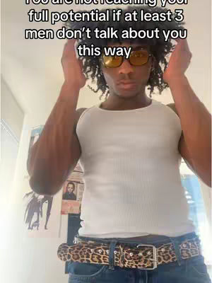 A young Black man stands indoors, facing the camera, with his palms up near his head. The background is a plain wall, and there are some posters. He wears a white tank top, blue jeans, and a leopard-print belt. The image has a simple, casual composition focused on the man and his pose.