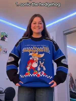 A young woman stands in front of what appears to be a bedroom. She is wearing a blue and black racing-style sweater featuring Sonic the Hedgehog and other characters from the franchise. The sweater is the main focus, and she smiles at the camera. The lighting suggests an indoor setting with blue LED lights on the wall behind her.