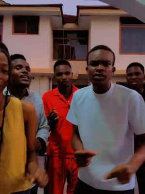A group of people, likely in a backyard or outdoor setting, are singing or possibly dancing. The focus is on a male in a white t-shirt leading the group, with others in various outfits. The backdrop includes a building.