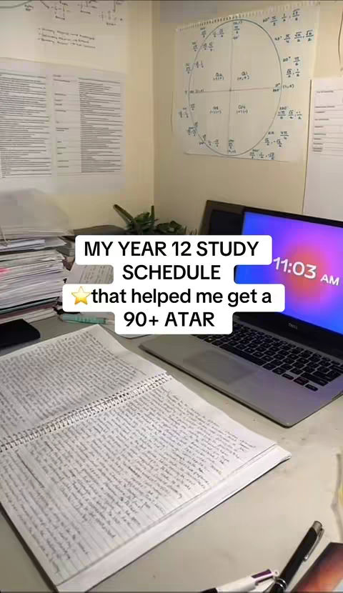 A shot of a desk with a laptop, notebook, and several sheets of paper. The laptop screen displays a purple and blue gradient with the time 11:03 AM. There is a notebook open with notes. The background includes a wall with notes and calculations, and a small plant.