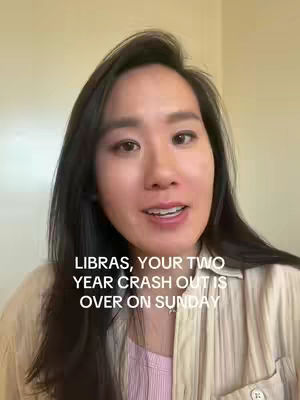 A woman with dark hair, wearing a light-colored shirt, is centered in the frame, looking directly at the camera and speaking. Text overlayed on the image reads, 'LIBRAS, YOUR TWO YEAR CRASH OUT IS OVER ON SUNDAY.' The background is a plain wall, and the lighting appears natural, suggesting an indoor setting.