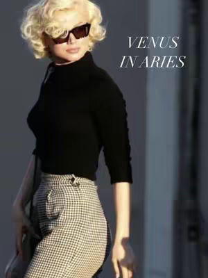 A woman with blonde curly hair wearing black sunglasses, a black top, and checkered pants poses with one hand on her hip, the words "VENUS IN ARIES" are in white text on the upper right of the frame.