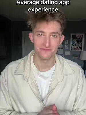 A young man with light skin and light brown hair is centered in the frame, looking towards the viewer with a subtle, slightly bemused expression. He is wearing a light beige jacket over a white t-shirt. The background is dimly lit, but shows some glimpse of an ambient room setting with shades of gray and a picture frame on the wall.