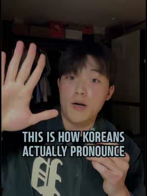A young man with short black hair is holding his hand out towards the camera, his fingers spread and his palm facing outwards. The background is a dimly lit room with items on shelves and what appears to be a closet door in the background. Text overlaid on the image reads, 'THIS IS HOW KOREANS ACTUALLY PRONOUNCE'.