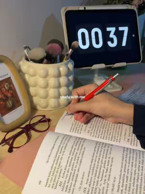 A photo of a well-lit desk with study materials. An open textbook with text is in focus. A red pen is poised above the page, as if in the act of writing. To the left sit purple glasses. Other visible items include a white container for makeup brushes, a digital clock showing '00 37' and a picture frame. The scene suggests a study environment.