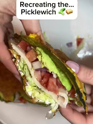 The image shows a close-up of a picklewich held between two hands against a blurred background. The sandwich features a dark-green exterior, likely a pickle, that encases various fillings including tomatoes with a shiny appearence, lettuce, sliced white meat. In the upper part of the image, a white text box sits over the cover images that says: Recreating the… and “Picklewich”with an emojy of cucumber and a sandwich. The person's hands are visible, one with pink nail polish.