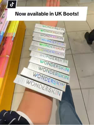 A person's arm, with many boxes of Wonderskin lip stain arranged on it. The arm is positioned with the palm facing the viewer, and the boxes are stacked up the forearm. The background includes a portion of a store shelf and the floor, implying a retail environment. A text overlay at the top reads "Now available in UK Boots!"