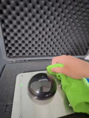 The scene presents a close-up view, the top part is a textured, dark gray surface with a honeycomb pattern. Below it, there is a metallic surface with a large, black, glossy, circular object in the center. A hand, holding a bright green cleaning cloth, appears from the right, actively cleaning the glossy black object. The light source seems to reflect off the objects.
