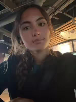 A selfie-style shot features a young woman in an indoor setting. The lighting is natural, with some light streaming in from the right side of the frame. She is wearing a black top. Her expression is neutral with a slight smirk, looking directly at the camera. The background is somewhat out of focus, suggesting an interior space, possibly a restaurant or public area, and there are some architectural elements visible, like a partial view of the ceiling with integrated lamps.