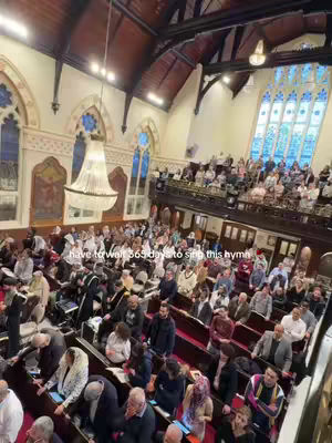 A wide shot of a large church interior filled with people. The church has high ceilings, ornate architectural details, and tall stained-glass windows. The image is well-lit, and the composition emphasizes the scale of the space and the multitude of individuals present.