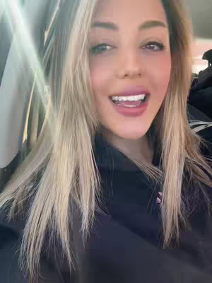 A medium shot captures a woman in the foreground, with a bright, well-lit background suggesting she might be in a car. She has long, slightly wavy blonde hair that frames her face and shoulders. The woman has a fair complexion, with a subtle application of makeup including pink lipstick. She is smiling with her mouth open, partially revealing her teeth, creating a friendly and inviting expression. The backdrop is blurred, suggesting movement or a shallow depth of field. The light is strong, with beams of light making the scene appear vibrant and cheerful.
