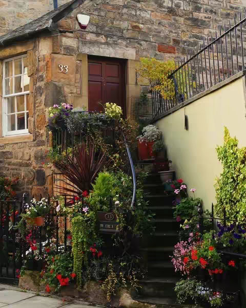 A stone building with a dark brown door, window, and a set of stairs leading up. The entrance and stairs are framed by lush greenery and flowers in pots of many colors. The stone exterior has various windows, and an address number is visible.