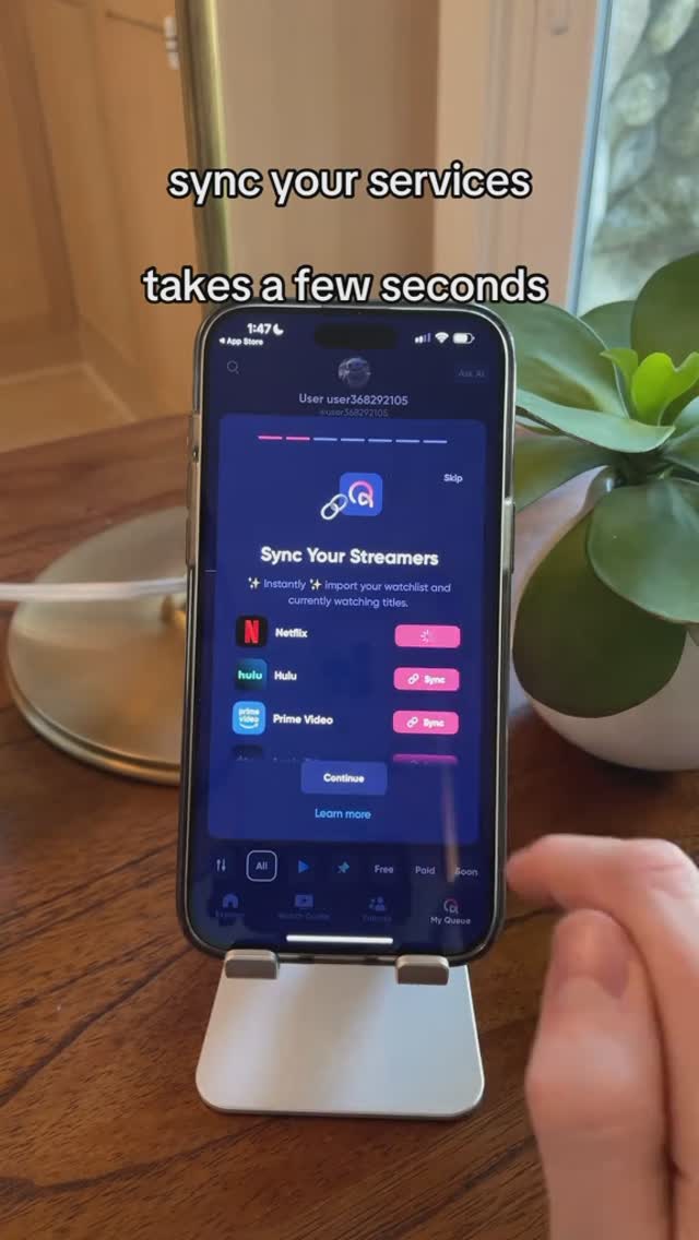A smartphone displaying an app screen is held by a person. The app interface is visible, featuring a blue and white theme with a highlighted section titled 'Sync Your Streamers.' Several streaming services, including Netflix, Hulu, and Prime Video, are listed with corresponding 'Sync' buttons. The phone is positioned on a stand resting on a wooden surface with a plant in the background