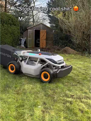 The image shows a robotic lawnmower in a grassy yard. In the background, there is a small wooden shed. The lawnmower has a gray and white body with orange wheels. The sky is overcast.