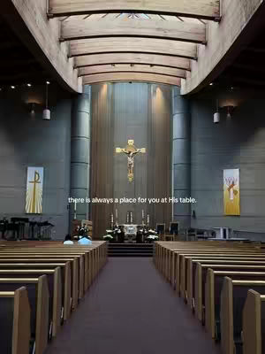 An interior shot of a church featuring rows of wooden pews leading to the altar. In the center, a crucifix is prominently displayed above the altar, with sunlight streaming in. The overall composition is symmetrical, emphasizing the architectural lines and the spiritual focus of the space.