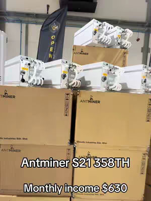 The image displays an indoor warehouse scene, likely a business or storage facility. The foreground is dominated by stacks of large, cardboard boxes labeled 'ANTMINER'. Several white, compact devices with attached cables and components are arranged on top of the boxes. In the background, a black flag bearing a logo or text is visible and provides context. The overall composition consists of the machines and boxes, stacked against industrial looking walls.