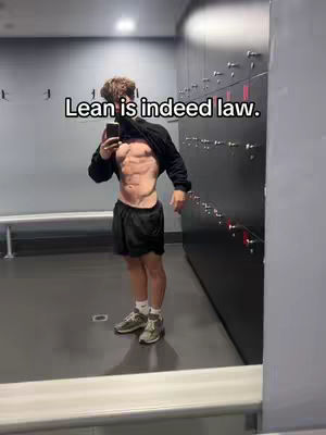 A muscular man stands in a gym locker room, taking a selfie in a mirror. He is shirtless, revealing his defined abs, and is wearing black shorts, sneakers, and a black hoodie over his shoulders. The image has an overlay of text that reads "Lean is indeed law."