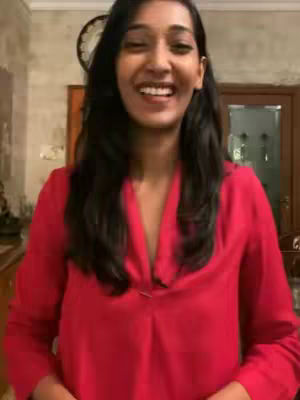 A smiling woman with long, dark hair wearing a red top is the focus. The background includes a clock and a door, suggesting an indoor setting. The lighting appears natural.