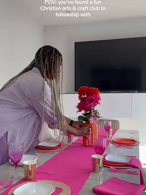 A woman with long, braided hair sets a table for a gathering. The table is dressed with a bright pink runner and matching napkins. Various place settings, including plates and glasses, are arranged alongside an orange vase filled with red flowers. The background shows a modern living space with a large television.