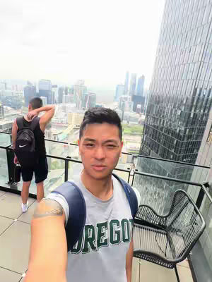 A selfie-style photo taken from a rooftop overlooking a cityscape with a modern skyscraper. The main subject is a man wearing a grey shirt with the word 'OREGON' printed on it. Another person is seen in the background. The lighting is bright and the composition emphasizes the urban setting.