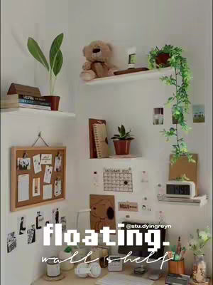 A well-lit, white-walled room with a desk setup. Floating white shelves hold plants, books, and decorative items. A bulletin board with notes and photos hangs on the wall. The overall aesthetic is clean and minimalist.