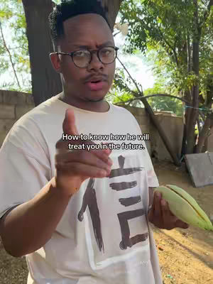 A man, wearing glasses and a white t-shirt, points towards the viewer while holding a green object. Behind him are trees and foliage.