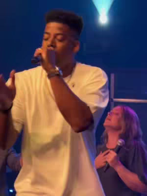 A live performance setting; a male singer holding a microphone takes center stage, dressed in a white t-shirt. Behind him, a woman with long hair appears to be worshipping, also holding a microphone. The lighting sets a blue background, accented by a spotlight above.