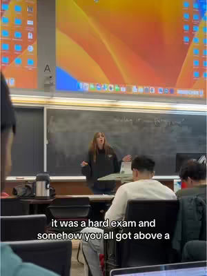 A woman stands in front of a chalkboard in a lecture hall, speaking to a class. Text overlays appear at the bottom of the image against the backdrop of the scene.