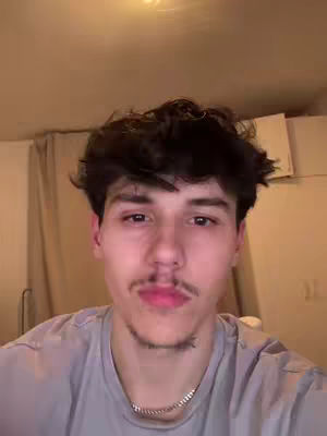 A selfie-style shot featuring a young man with dark hair and a slight mustache, centered in the frame. He's wearing a light grey t-shirt and a silver chain necklace. The background is dimly lit, suggesting an indoor setting with curtains and a ceiling light visible. The lighting is soft and diffused.