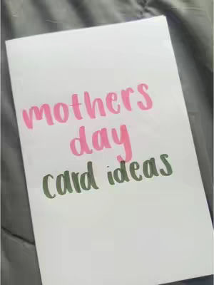 A white card is angled towards the viewer, upon which there is a hand-written header; the card sits upon a textured fabric that appears to be a gray comforter or blanket. The text is in a handwritten font. The top line of text is pink and reads “mothers”. The second line of text is pink and reads “day”. The third line of text is green and reads “card ideas”.