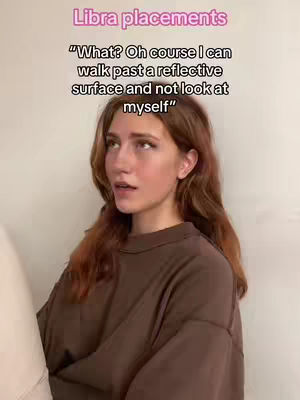 A young woman with auburn hair is positioned slightly off-center, looking towards the left. Above her, the image features text in a soft pink hue: 'Libra placements'. Below the text, a humorous quote is displayed in black: 'What? Oh course I can walk past a reflective surface and not look at myself'. The background consists of a solid, off-white wall. The woman wears a loose-fitting, brown crew-neck shirt.