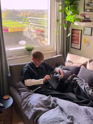 A young man is lying on a bed or couch in a sunlit room, reading using a tablet. The room has large windows with a view of the outdoors and some plants hanging on the wall.