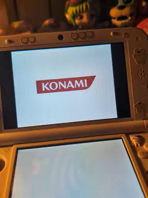 Image shows a white Nintendo 3DS console, with a screen displaying the KONAMI logo in red on a white background. The console is in focus, and the background is blurred but appears to be a room. The image is lit, possibly softly, and the composition focuses on the game screen.
